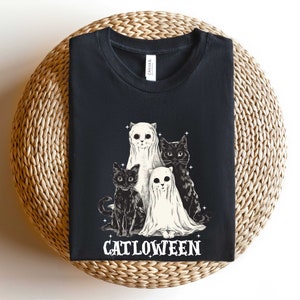K&ouml;nnte beinhalten: Schwarzes T-Shirt mit einem Aufdruck von drei Katzen, die als Geister verkleidet sind. Der Text "Catlowteen" ist unter den Katzen aufgedruckt.