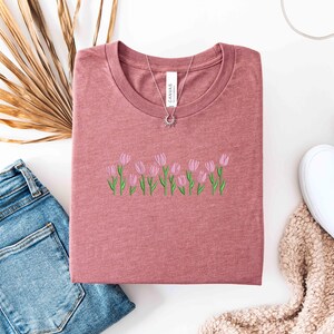 K&ouml;nnte beinhalten: Staubrosafarbenes T-Shirt mit Rundhalsausschnitt, bestickt mit rosa Tulpen und gr&uuml;nen Stielen. Eine silberne Halskette mit Sonnenanh&auml;nger liegt auf dem Shirt. Das Shirt liegt neben einer blauen Jeans und wei&szlig;en Turnschuhen.