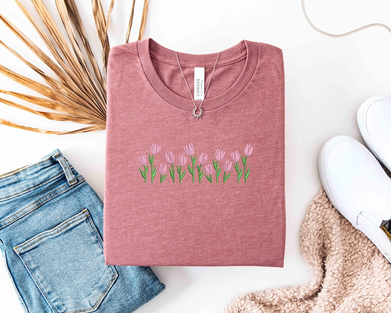 K&ouml;nnte beinhalten: Staubrosafarbenes T-Shirt mit Rundhalsausschnitt, bestickt mit rosa Tulpen und gr&uuml;nen Stielen. Eine silberne Halskette mit Sonnenanh&auml;nger liegt auf dem Shirt. Das Shirt liegt neben einer blauen Jeans und wei&szlig;en Turnschuhen.