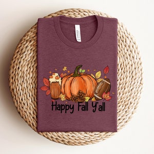 May include: A maroon t-shirt with a graphic of a pumpkin, a football, and a glass of beer with the text "Happy Fall Y'all".