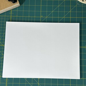 May include: A white sheet of paper on a green cutting mat with yellow grid lines. There is a white ruler and a small piece of brown fabric on the mat.