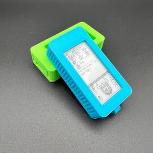 May include: A handheld electronic device with a screen displaying text and graphics, encased in a bright blue protective case. The screen shows the word "BJORT", and the text "You say hack, I say how high?" A green case sits behind it.