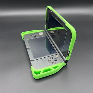 May include: A gray handheld gaming console with a bright green protective case. The console is open, revealing two screens and several buttons. The case wraps around the edges of the device, providing a contrasting color accent.