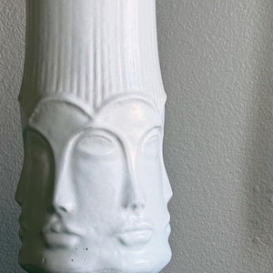 Può includere: Un vaso in ceramica bianca con un design unico che presenta più volti. Il vaso ha una forma cilindrica con scanalature verticali e un motivo di volto dettagliato. L'altezza è di circa 30 cm.