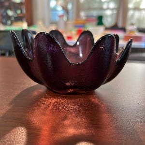 Può includere: Una ciotola di vetro viola scuro con un design unico a forma di petalo. La ciotola ha una superficie strutturata ed è appoggiata su un tavolo di legno. La forma della ciotola ricorda un fiore, con bordi curvi e un colore scuro e ricco.