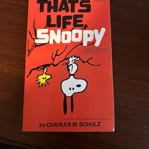 May include: A vintage paperback book titled "That's Life, Snoopy" with a red cover. The cover features a cartoon illustration of Snoopy and a yellow bird. The book is by Charles M. Schulz.