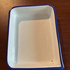 May include: A rectangular white enamel tray with a blue border. The tray has a slightly curved shape and a smooth, glossy surface. The blue border adds a contrasting accent to the white interior.