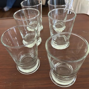 May include: A set of six clear glass tumblers arranged in a group. Each tumbler features a smooth, cylindrical body that tapers slightly toward a reinforced, circular base. The glass is transparent with no patterns or colors, showcasing a simple design.