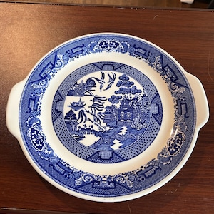 May include: White and blue ceramic serving platter with a Willow pattern design. The platter has a round shape with two handles and a detailed blue pattern around the rim and center. The center features a scenic landscape.