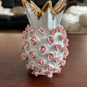 May include: A white ceramic pineapple-shaped vase with gold accents and pink rose embellishments. The pineapple crown is trimmed in gold. The vase is a decorative item.