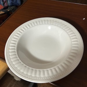 May include: A round white ceramic bowl with a wide, flat rim featuring a decorative embossed vertical ribbed pattern and a beaded edge detail. The bowl has a smooth, glossy finish and a deep, concave center for serving food.