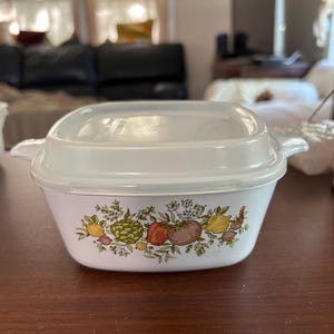 May include: White rectangular casserole dish with a clear lid. The dish features a colorful border of various vegetables, including tomatoes, lemons, and artichokes. The dish has two small handles on either side.