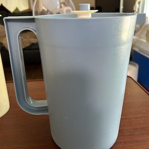 May include: A light blue plastic pitcher with a sturdy side handle and a matching lid featuring a small, centered white knob. The cylindrical container has a smooth finish and a simple, functional design with a pouring spout on the upper rim.