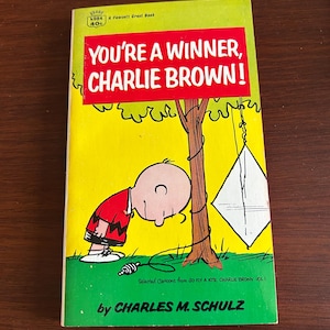 May include: A vintage paperback book titled "You're a Winner, Charlie Brown!" featuring a cartoon illustration of Charlie Brown with a kite. The cover is yellow and green with red text. The book is by Charles M. Schulz.