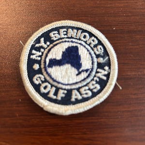 May include: Circular patch with the text "N.Y. SENIORS GOLF ASS'N" in white lettering on a navy blue background. The center features a white circle with a blue outline of New York State.