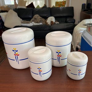 May include: A set of four white ceramic canisters of varying sizes, each featuring a horizontal blue line and a small, stylized floral motif in yellow, red, and blue. Each canister includes a matching white lid with a recessed handle.