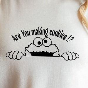 Könnte beinhalten: Cremefarbenes T-Shirt mit einer schwarzen Grafik eines Cartoon-Monsters, das über den unteren Rand lugt. Der Text über dem Monster lautet "Are You making cookies !?" in einer verspielten Schriftart. Es ist ein lässiges Kleidungsstück für den Alltag.