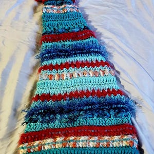 May include: A crocheted mermaid tail blanket in shades of teal, blue, and red. The tail is made with a variety of stitches and textures, including fringe and bobbles.