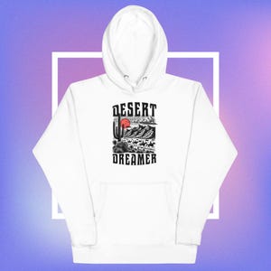 May include: White hoodie with a front graphic featuring a desert landscape with a cactus, mountains, and a red sun. The text "DESERT DREAMER" is printed above and below the graphic in a bold font. The hoodie has a drawstring hood and a front pocket.
