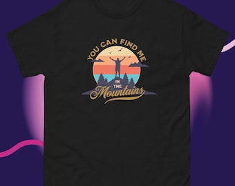 May include: Black t-shirt with a graphic design featuring a mountain scene and the text "YOU CAN FIND ME IN THE Mountains". The design includes a person with arms raised, a sunset, and birds.