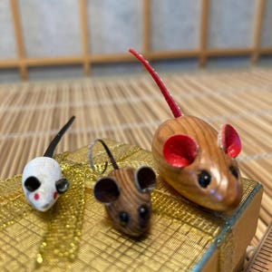 Puede incluir: Tres figuras de ratones de madera en miniatura. Uno es blanco con orejas negras y cola negra, otro es marrón con orejas negras, y el tercero es marrón con orejas rojas y cola roja. Los ratones se exhiben sobre una superficie dorada.