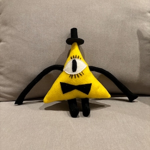 May include: A handmade yellow triangle plush toy with a black top hat, bow tie, and arms. The toy has a single eye with a white iris and black pupil. The plush is a character from the animated series Gravity Falls.
