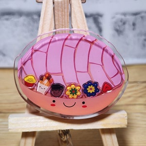Può includere: Un ciondolo in resina rosa e arancione a forma di concha, un pane dolce messicano tradizionale, con un viso sorridente e decorazioni di cibo di cartoni animati colorati. Il ciondolo è attaccato a un portabadge retrattile.