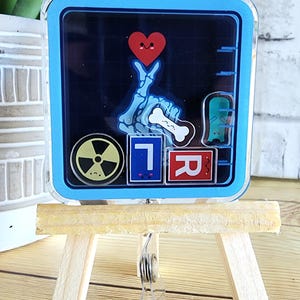 Puede incluir: Un soporte para credenciales cuadrado azul y negro con una mano de esqueleto haciendo un signo de paz, un corazón rojo, un hueso, un fantasma, un símbolo de radiación y tres bloques con las letras "T", "I" y "R" en un caballete de madera.