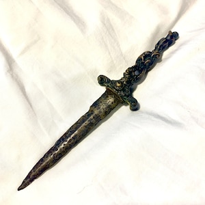 Może przedstawiać: Dekoracyjny sztylet z zakręconym, ozdobnym uchwytem i długim, ostrym ostrzem. Uchwyt wykonany jest z metalu i ma ciemne, postarzane wykończenie. Ostrze również jest wykonane z metalu i ma matowe, srebrne wykończenie.