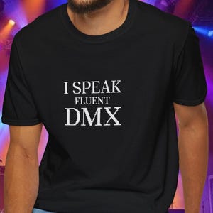 Może przedstawiać: Czarny t-shirt z białym napisem "I SPEAK FLUENT DMX". Osoba na zdjęciu ma na sobie niebieskie dżinsy. Tło przedstawia oświetlenie sceniczne.