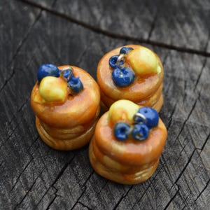 May include: Three miniature pancake charms, each featuring a stack of golden-brown pancakes topped with a yellow pat of butter and several small, glossy blue berries. Each charm includes a small silver-toned metal loop at the top for attachment.
