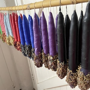 Puede incluir: Una fila de velas cónicas en un espectro de colores que incluye blanco, amarillo, verde, rosa, azul, morado y negro, colgadas de una varilla de madera. Cada vela está cubierta en la base con una capa gruesa de hierbas secas y flores.