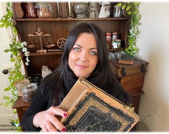 May include: A woman holding two antique books with ornate gold detailing. The books are aged with worn covers. A wooden cabinet with various items, including jars, scales, and a pentagram, is in the background. The scene has a vintage, mystical theme.