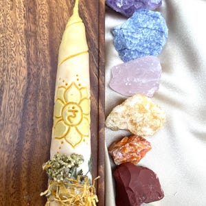 Puede incluir: Una vela de color crema con un diseño floral amarillo y flores secas en la base. Varios cristales en bruto coloridos, incluyendo amatista, calcita azul, cuarzo rosa y cornalina, están dispuestos junto a la vela sobre una superficie satinada.