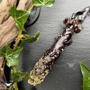 May include: A dark purple wand with a textured surface, decorated with red berries and green leaves. The wand's handle is embellished with a gold-coloured substance. The image includes green ivy and a piece of driftwood, suggesting a mystical theme.