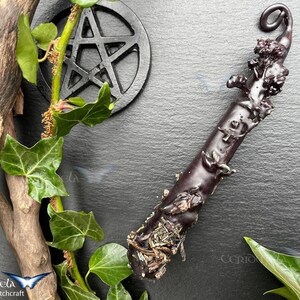 Puede incluir: Una varita de madera negra tallada a mano con un diseño en espiral y un pequeño detalle de hojas en la parte superior. La varita está sobre una superficie negra con hojas de hiedra verdes.