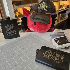 May include: Two black metal hip flasks with a gold design featuring a creature playing a guitar and the text "Bardic Inspiration A celebration of the magic in music". One flask is on a gray cutting mat, the other is on a white surface. A camouflage baseball cap with a yellow and green creature graphic is on a red plush toy.