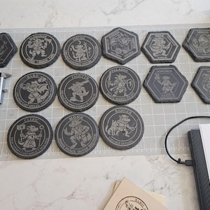 以下が含まれることがあります： ファンタジーキャラクターのデザインが刻まれた、濃い灰色のスレートコースターのコレクション。コースターは丸型と六角形で、バーバリアン、ドルイド、レンジャーなどのキャラクターが描かれています。左側にはデジタルノギスが見えます。