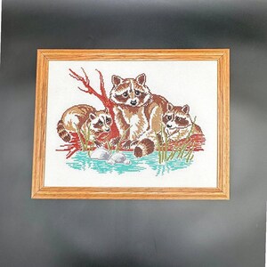 May include: Cross-stitch artwork of three raccoons in a natural setting, framed in light brown wood. The raccoons are depicted in shades of brown and tan, with a backdrop of green grass, blue water, and a red tree branch. The artwork is on a white background.