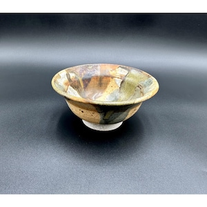 May include: A handmade ceramic bowl with a wide, rounded shape. The bowl features a speckled, earthy color palette with shades of brown, tan, green, and gray. The interior has a glossy finish, while the exterior has a textured appearance. The bowl sits on a small, unglazed base.