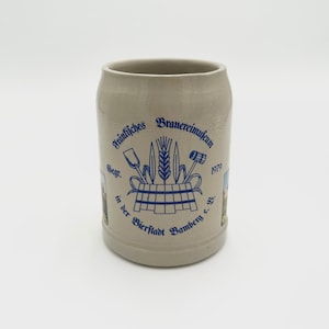 May include: A beige ceramic beer stein with a blue and white logo that reads "Traditionelles Brauereimuseum 1979 Gegr. in der Bierstadt Bamberg e.V."