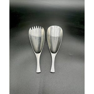 May include: Two silver serving spoons on a black background. One spoon has a slotted, fork-like end, while the other has a smooth, oval bowl. Both spoons have a slender handle.