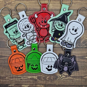 May include: Set of ten Halloween-themed keychains featuring various characters, including a mummy, a ghost, a witch, a devil, a Frankenstein monster, a bat, and three buckets. The keychains are made of felt and have embroidered details.