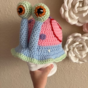 May include: A crochet bucket hat designed to resemble a snail, featuring a light blue base with a light green scalloped brim. The top has a pink shell pattern with red accents and two tall, light blue stalks topped with light green eyes.