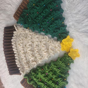 May include: Crocheted Christmas tree ornaments in green, cream, and brown. The green trees are decorated with colorful beads. Two yellow star ornaments are also present. The ornaments are on a white, fluffy surface.