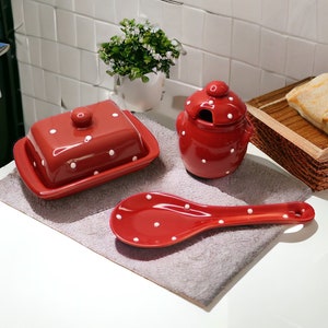 May include: Red and white polka dot ceramic kitchenware set including a butter dish, sugar bowl with lid, and a spoon.