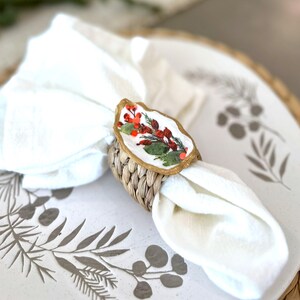 May include: A white linen napkin is held by a woven napkin ring with a gold-trimmed, oval-shaped centerpiece. The centerpiece features a floral design with red berries and green leaves. The napkin and ring rest on a white plate with a botanical pattern.
