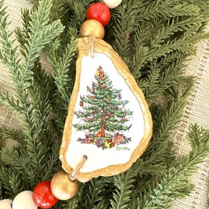 May include: A Christmas ornament with a painted Christmas tree design, attached to a beaded garland. The garland features white, red, and gold wooden beads. The ornament is in the shape of an oyster shell. The word "Spode" is printed on the ornament.