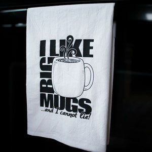 May include: White kitchen towel with black text and a coffee mug graphic. The text reads "I LIKE BIG MUGS...and I cannot lie!" The mug has steam rising from it. The towel is hanging in front of a dark background.