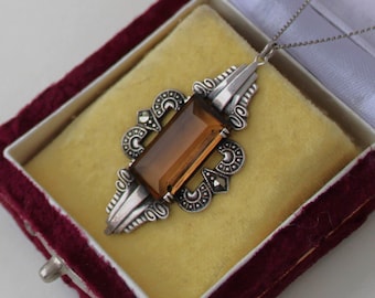 May include: A silver pendant necklace with a rectangular amber-colored gemstone set in a detailed Art Deco design. The pendant is in a velvet jewelry box.